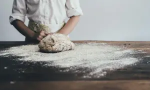 Ciasto na drewnianym stole, posypane mąką, w tle osoba w fartuchu.