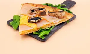 Kawałek pizzy capriciosa z szynką, serem i pieczarkami na ciemnej desce.