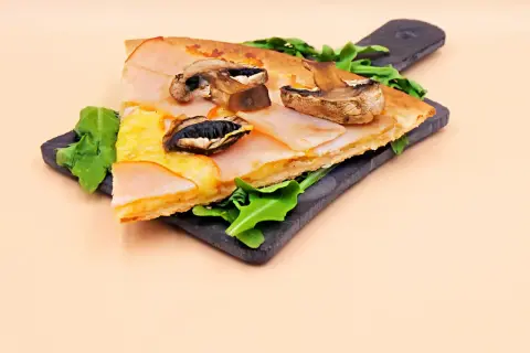 Kawałek pizzy capriciosa z szynką, serem i pieczarkami na ciemnej desce.