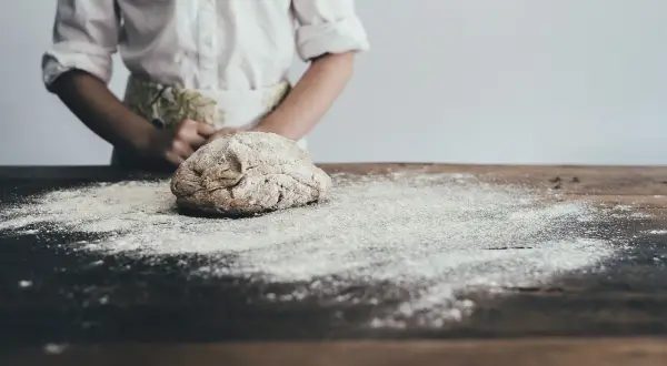 Ciasto na drewnianym stole, posypane mąką, w tle osoba w fartuchu.